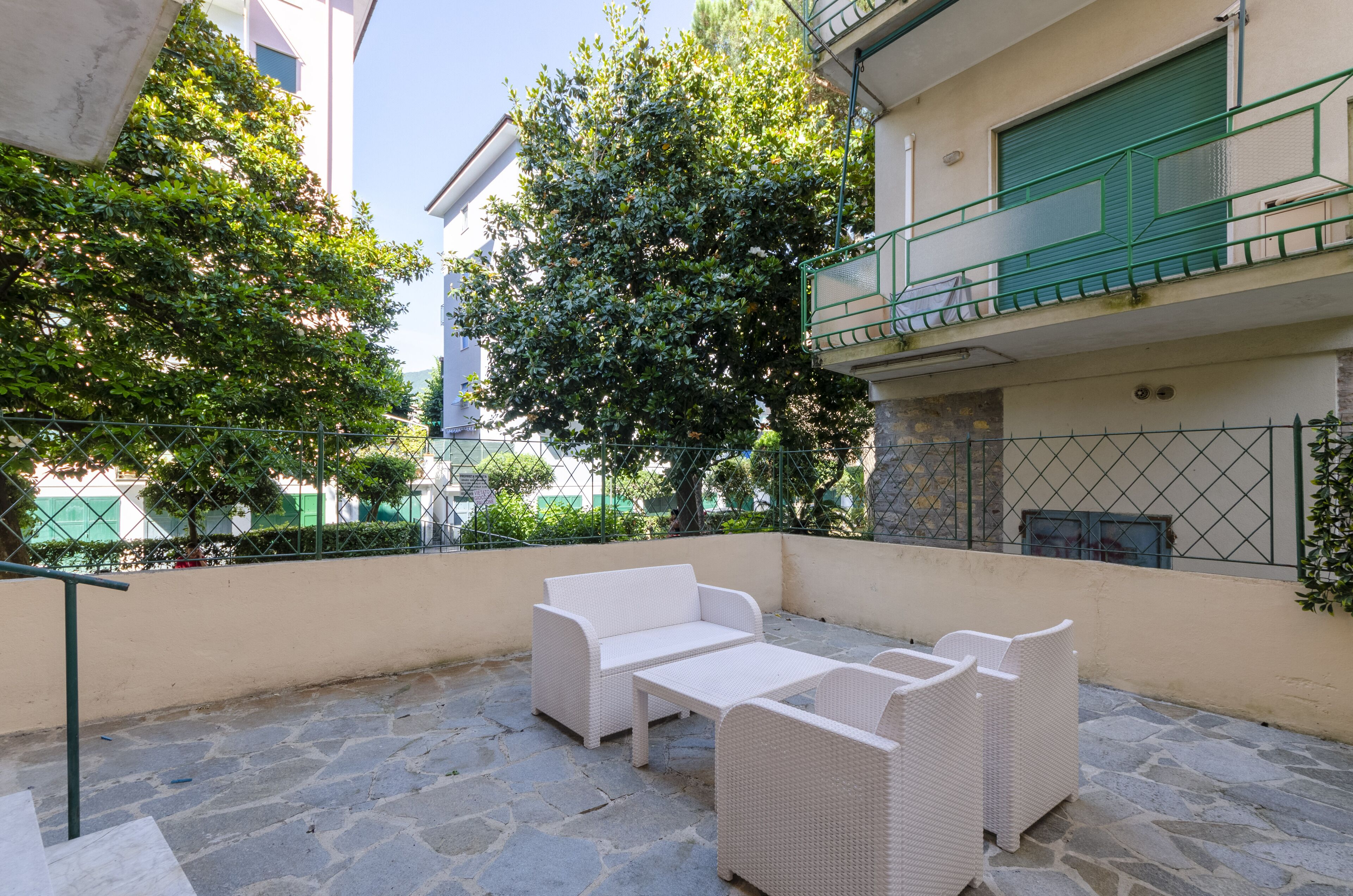An outdoor patio with a white wicker sofa set and a table, surrounded by stone flooring and greenery.