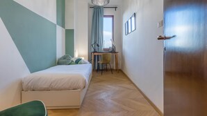 Ein Schlafzimmer mit Bett, Schreibtisch und Stuhl. Rechts ist eine Tür und ein Fenster mit Vorhängen.