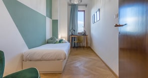 A bedroom with a bed, a desk, and a chair. There is a door on the right and a window with curtains.