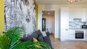 Eine moderne Küche mit weißen Schränken und einer Marmorarbeitsplatte, ein graues Sofa mit Kissen sowie eine Wand mit einem tropischen Pflanzenwandbild.