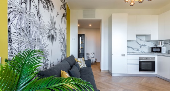 A modern kitchen with white cabinets and a marble countertop, a gray sofa with cushions, and a wall with a tropical plant mural.