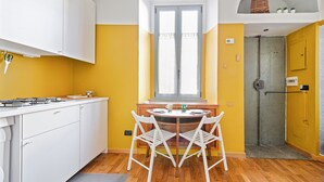 A kitchen with white cabinets, a wooden dining table with two white chairs, a window with curtains, and a door leading to a bathroom. - JOIVY Bright And Cozy Studio For 2 In Porta Genova (Milan)