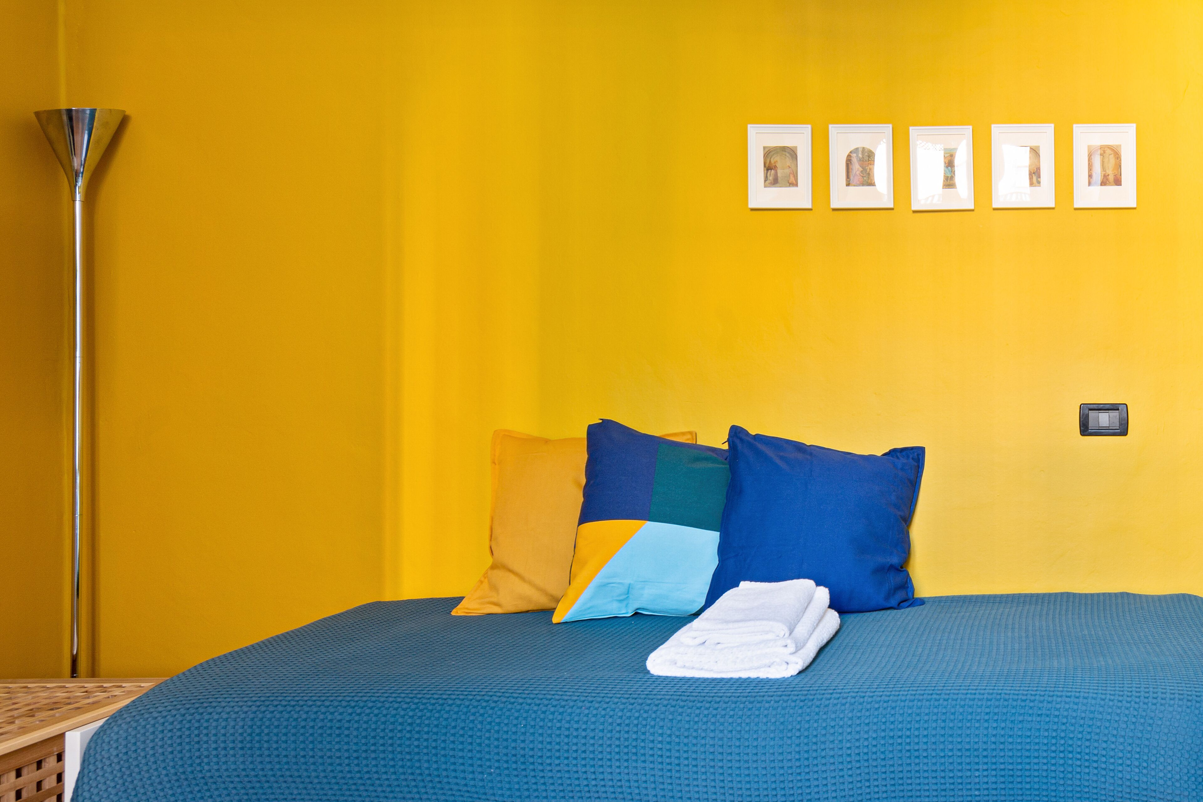 A room with a bed, pillows, a folded towel, and a wall with framed pictures.