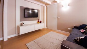 Un salon moderne avec un téléviseur à écran plat fixé au mur, une console blanche agrémentée d’objets décoratifs, un tapis beige et un canapé recouvert d’un plaid à motifs floraux.