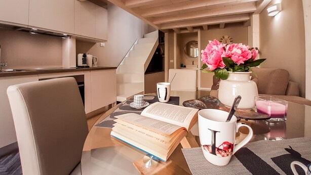 A modern kitchen with a dining area, featuring a wooden table with a book, a mug, and a vase with flowers.