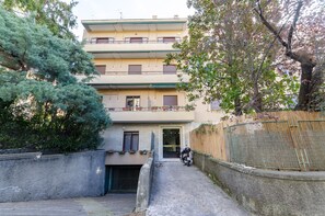 Ein mehrstöckiges Wohnhaus mit geschlossenem Eingangsbereich, einem Motorrad vor der Tür und einem Baum im Vordergrund.