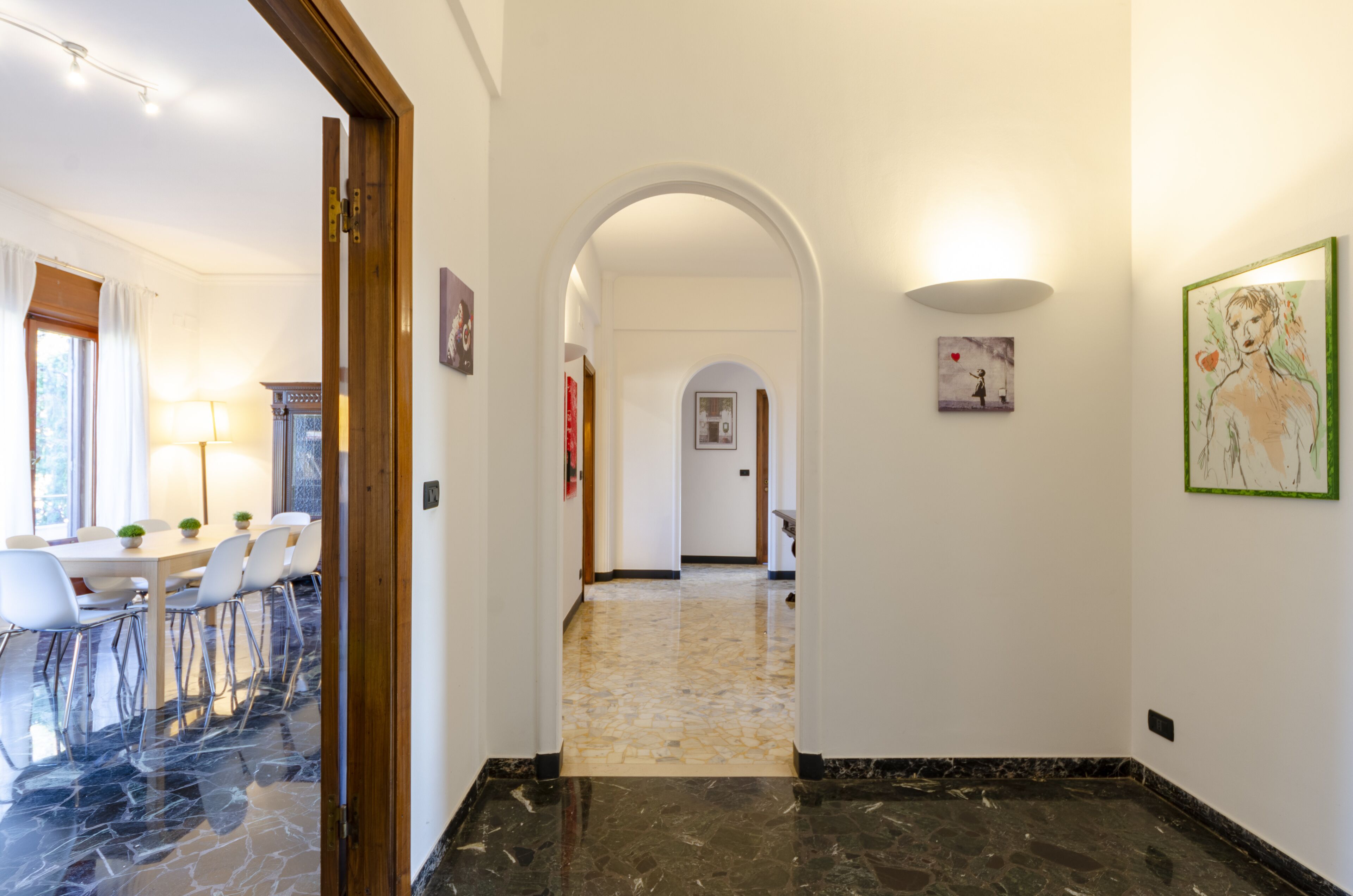 A hallway with a marble floor, white walls, and an archway leading to another room. There are framed artworks on the walls and a dining area with chairs and a table visible on the left.
