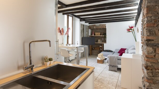 A modern kitchen with a stainless steel sink, wooden countertop, and a dining area with a white table and chairs. There is a brick wall and a sofa with a striped throw blanket in the living area. - JOIVY Cosy Apt in Como Centre (Como)