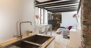 Cozinha moderna com pia de aço inoxidável, bancada de madeira e espaço de refeições com mesa e cadeiras brancas. Há uma parede de tijolos e um sofá com manta listrada na sala de estar.