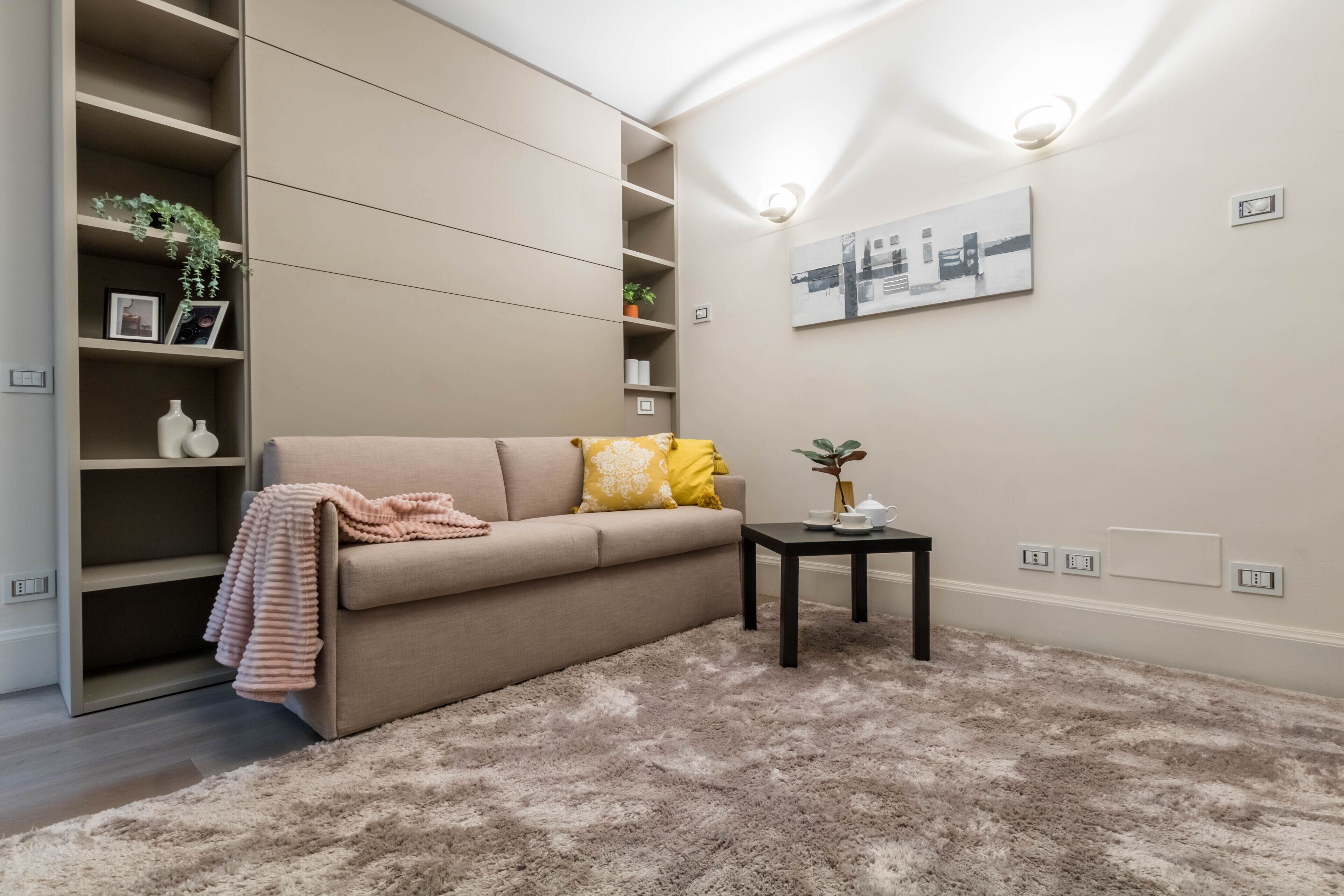 Un salon avec un canapé beige, une petite table d’appoint, un tapis moelleux et des étagères intégrées où sont exposés des objets décoratifs.