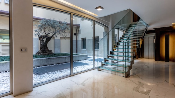 Un intérieur moderne avec un escalier en verre menant à un deuxième étage, une grande porte en verre offrant une vue sur un arbre et une cour, et un sol en marbre.