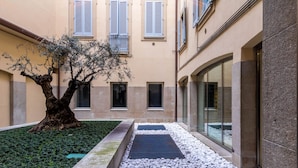 Une cour avec un arbre, un chemin et des bâtiments dotés de fenêtres et de volets.