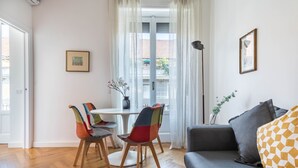 A modern living room with a grey sofa, colorful chairs, a dining table, and a large window with curtains.