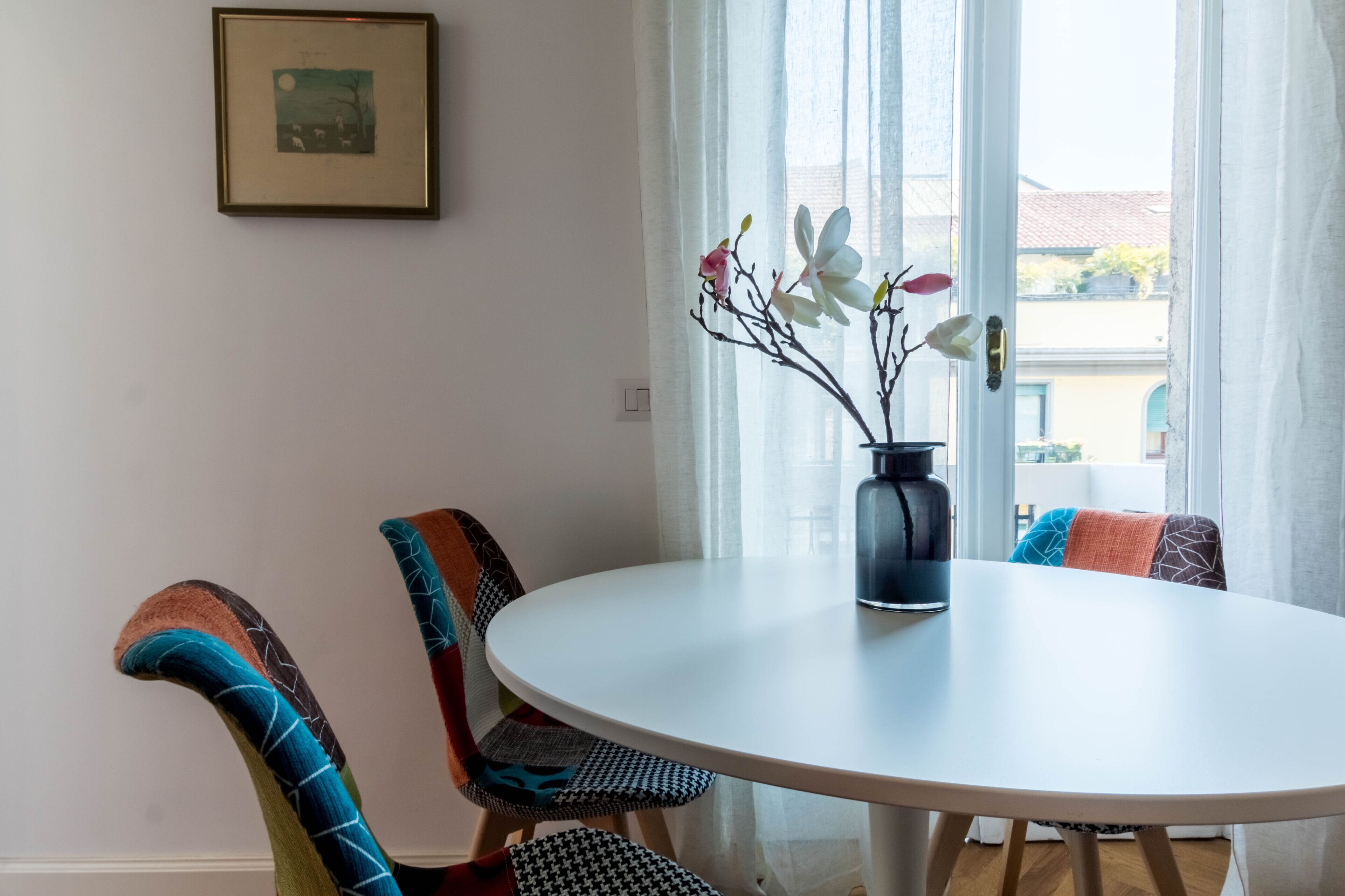 A round dining table with a vase of flowers, two chairs with patterned upholstery, and a framed picture on the wall.