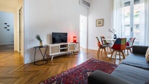 A modern living room with a flat-screen TV, a dining area with colorful chairs, and a large window with sheer curtains.