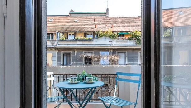 A balcony with a table and chairs, a view of buildings with red-tiled roofs, and a window frame.