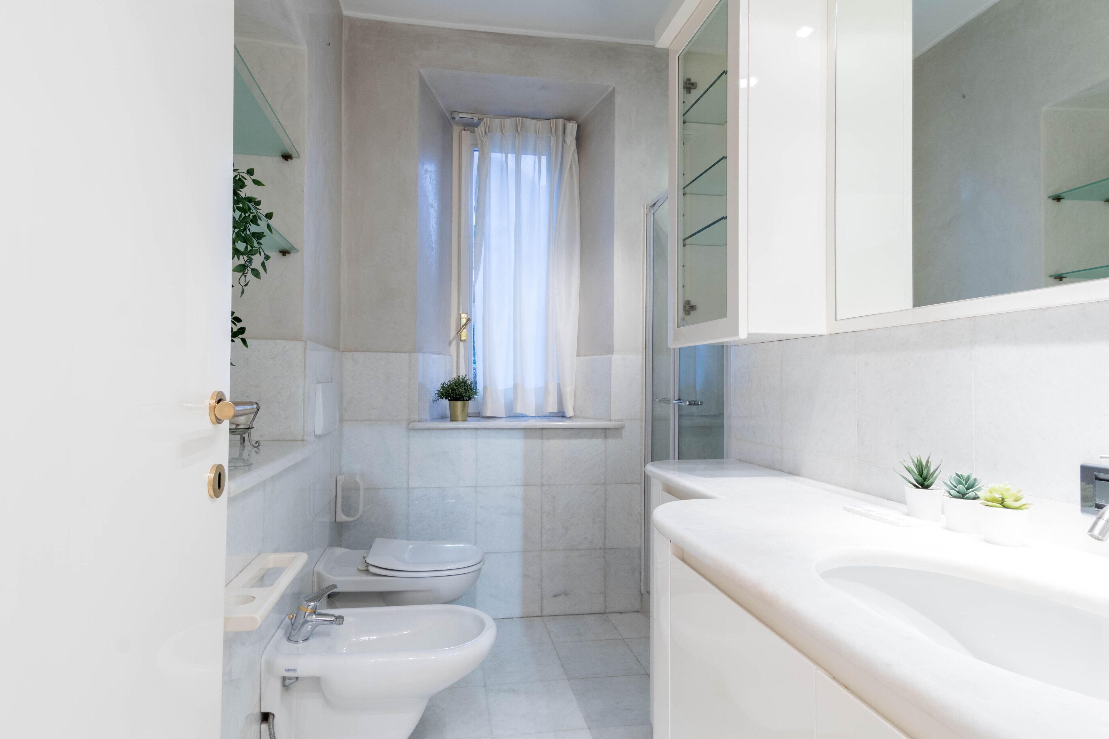 A modern bathroom with a bathtub, a toilet, and a bidet. There is a window with a curtain and a small plant on the windowsill.