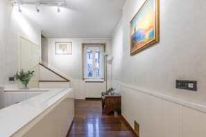 A hallway with a wooden floor, white walls, a staircase, a window with a view, and a painting on the wall.