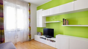 A modern living room with a flat-screen TV on a white entertainment unit, a bookshelf with books and decorative items, and a window with sheer curtains. - JOIVY Lovely Flat For 4 W/Balcony Near Porta Romana (Milan)