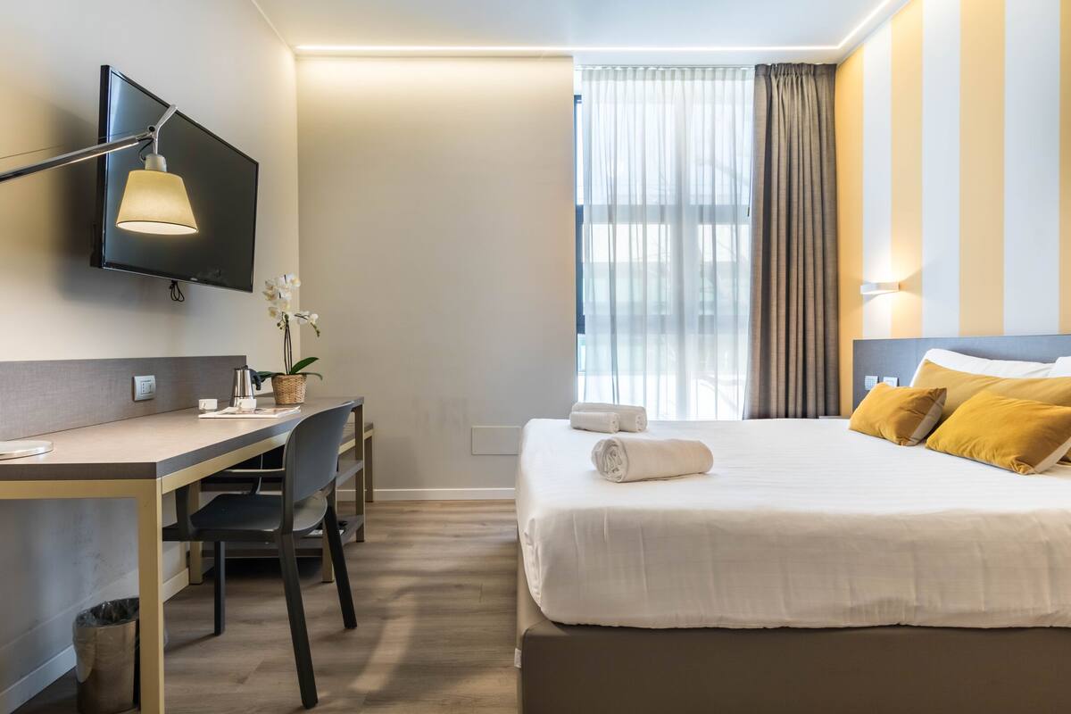 A hotel room with a bed, a desk, a chair, a TV, and a window with curtains.
