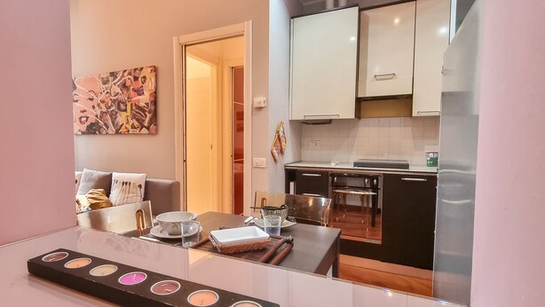 Una cucina moderna con mobili bianchi, un tavolo da pranzo con piano in vetro e un quadro astratto dai colori vivaci appeso al muro.