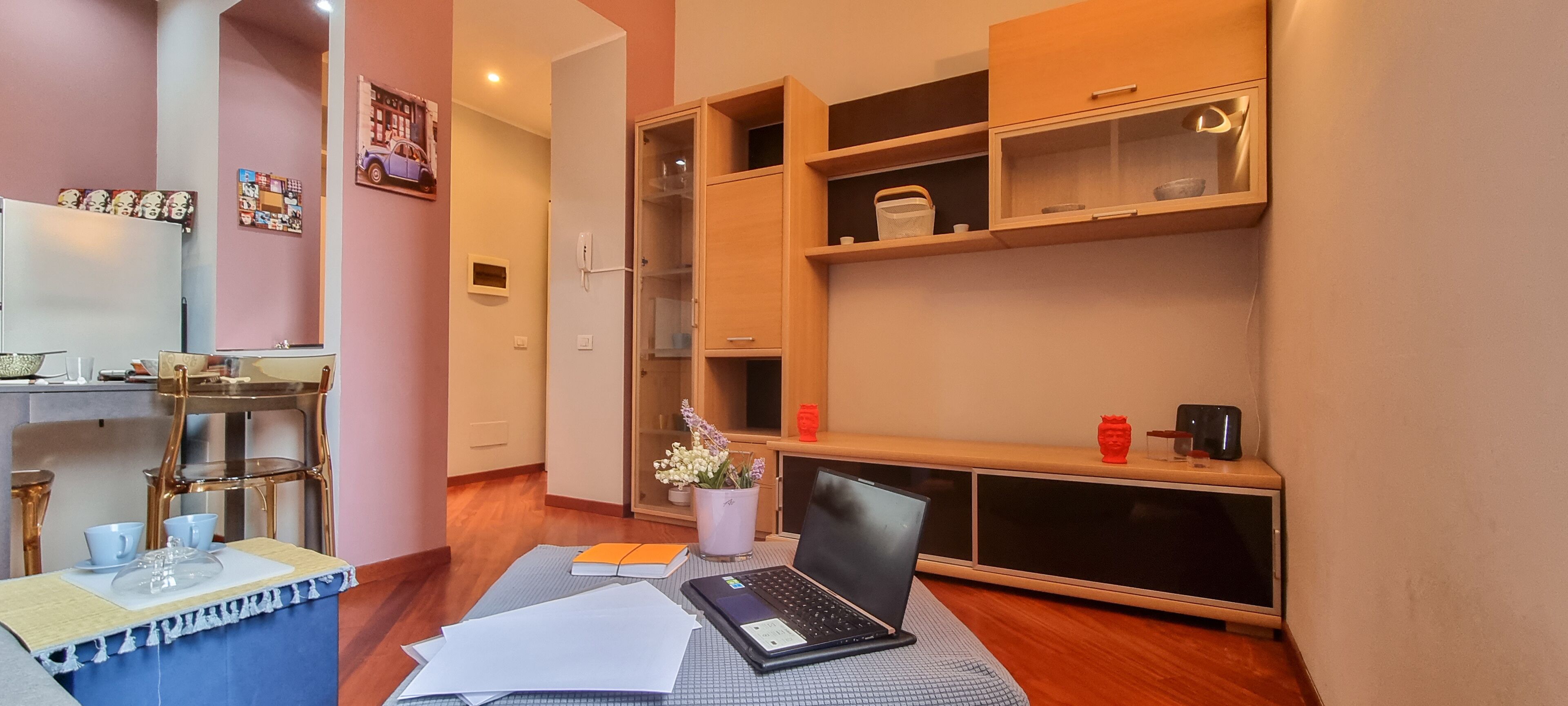 A modern room with a desk, laptop, and papers. There is a bookshelf with decorative items and a small table with a vase of flowers.