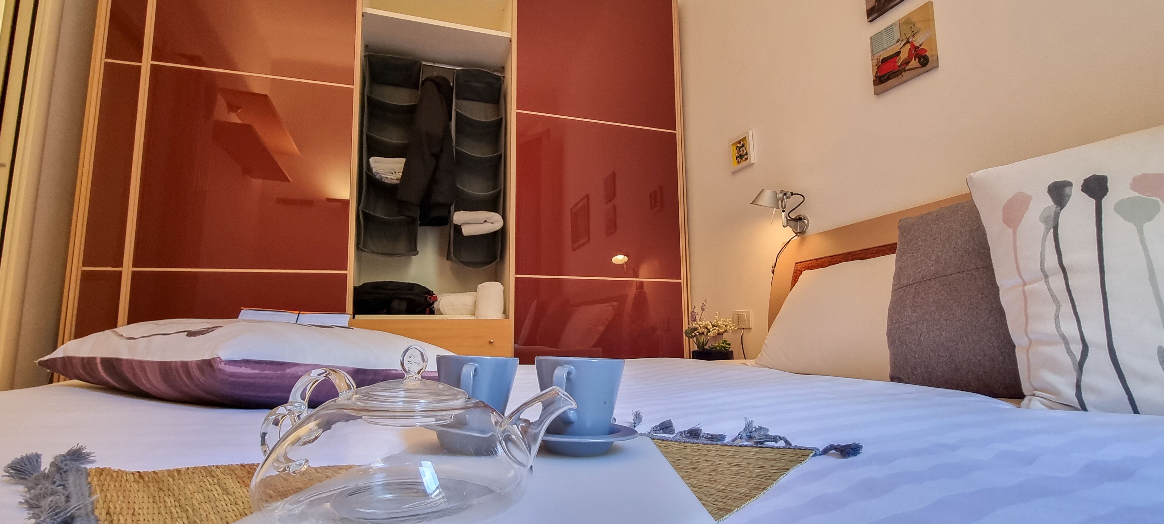 A hotel room with a bed, a wardrobe, a bedside table with a teapot and cups, and a nightstand with a lamp.