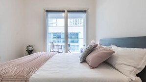 A bedroom with a bed, a chair, a small table, and a window with a view of buildings.
