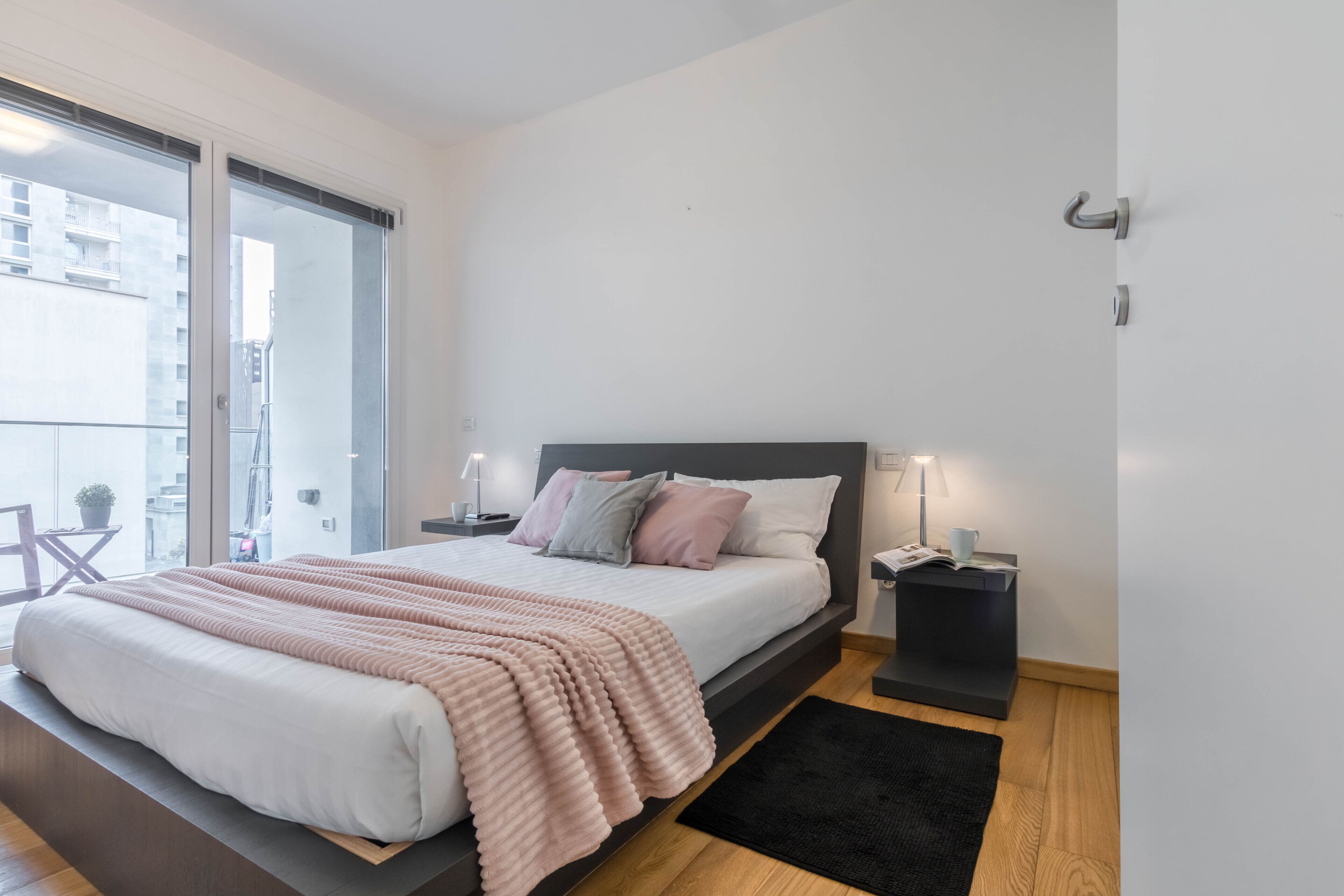 A modern bedroom with a large bed, bedside tables, a black rug, and a view of the city through a sliding door.
