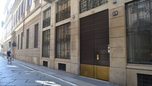 Une rue bordée d’un immeuble doté de grandes fenêtres avec des grilles en métal, d’une enseigne portant le numéro « 19 » et d’un cycliste qui passe.