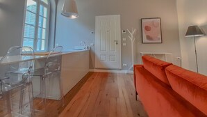 Une pièce avec un plancher en bois, une porte blanche, une table en verre transparent avec des chaises, un canapé rouge et une lampe.
