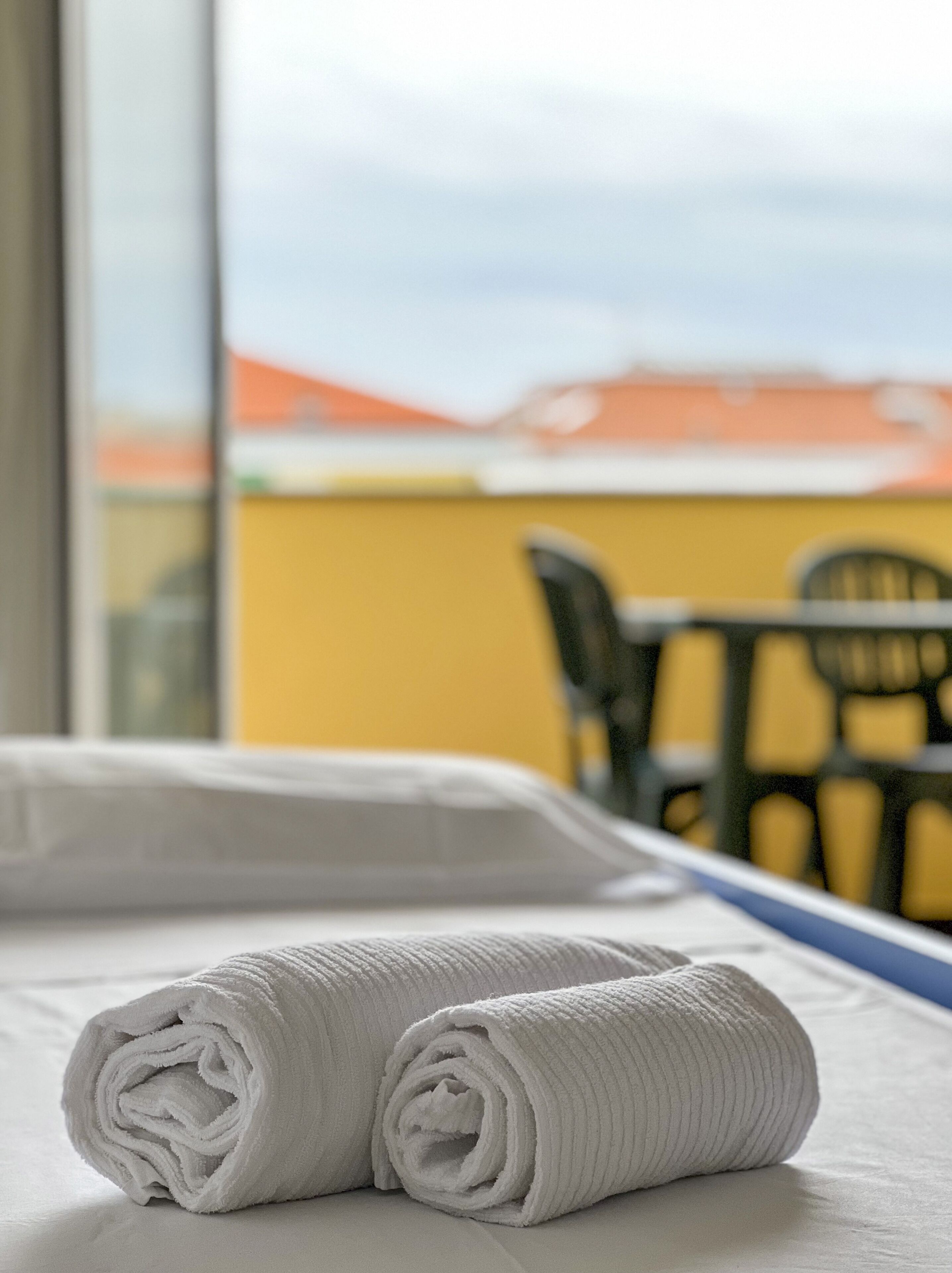 A neatly made bed with rolled white towels, a window with a view of buildings, and a table with chairs outside.