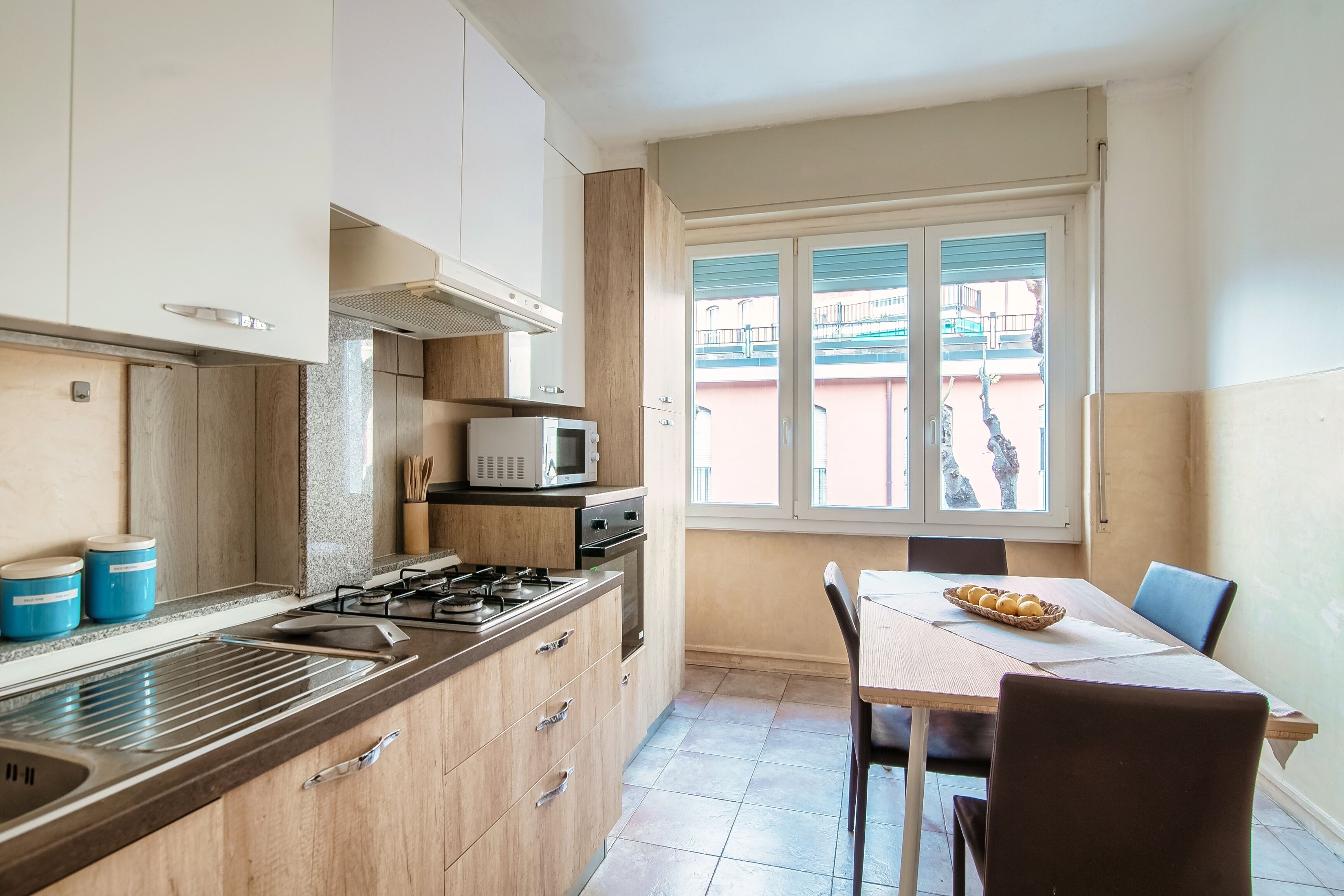 A compact kitchen with a gas stove, microwave, and wooden cabinets. A dining table with chairs is positioned near a window with a view of buildings.