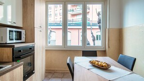 Ett kompakt kök med mikrovågsugn, ugn och skåp. Ett matbord med en korg full av citroner syns. Ett fönster med utsikt över en byggnad och en staty finns också.