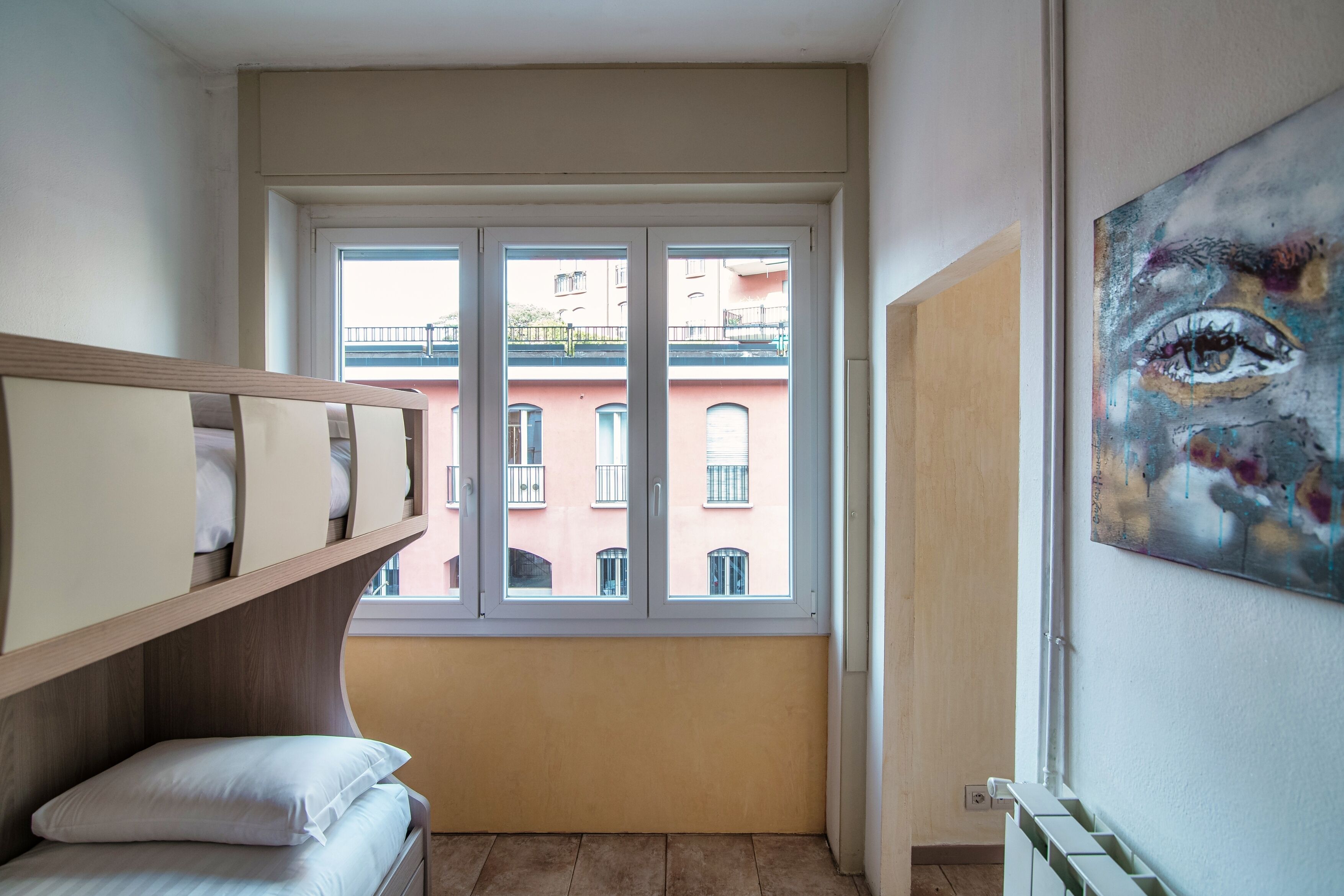 A room with a bunk bed, a window with a view of buildings, and a painting on the wall.