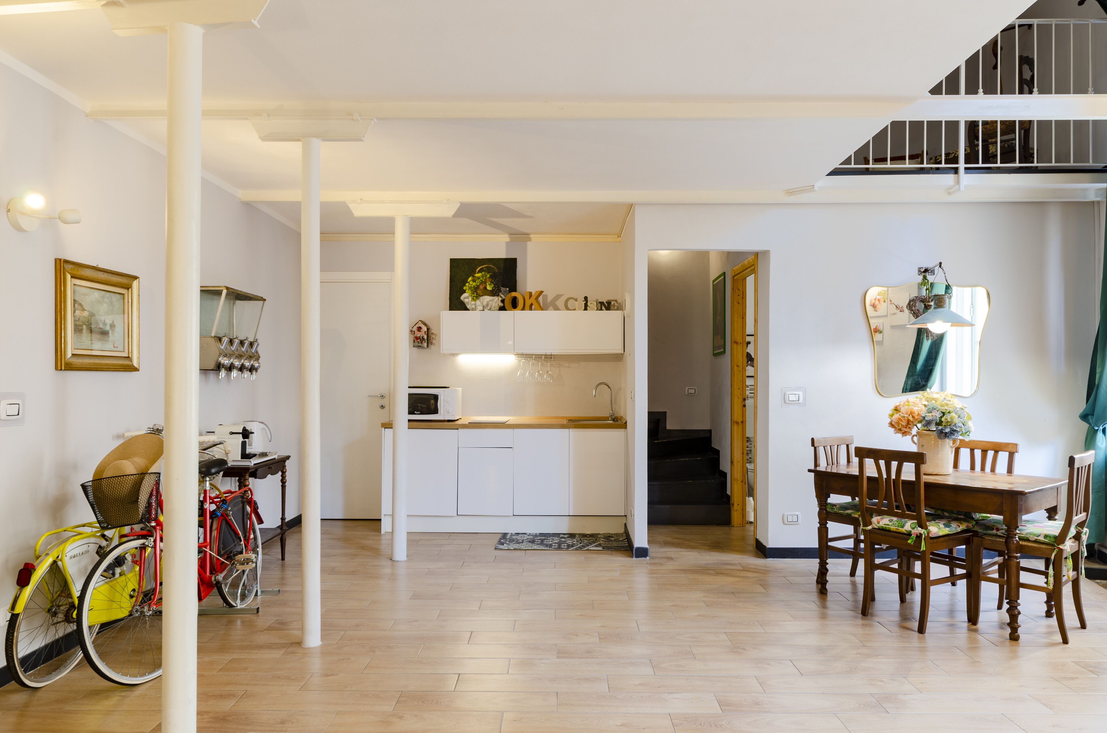 A spacious living area with a wooden floor, a dining table with chairs, a kitchen area with white cabinets, and a bicycle in the corner.