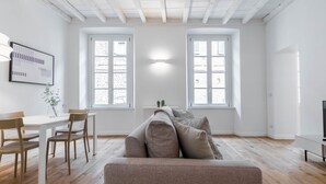 A modern living room with a sofa, dining table, and chairs. There are two windows with white frames and a ceiling with exposed beams.