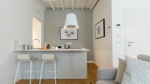 A modern kitchen with a central island, two bar stools, a hanging light fixture, and a grey sofa with patterned cushions.