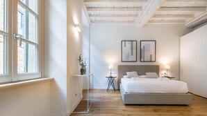 A bedroom with a large bed, two bedside tables with lamps, a small side table, and a window with blinds.