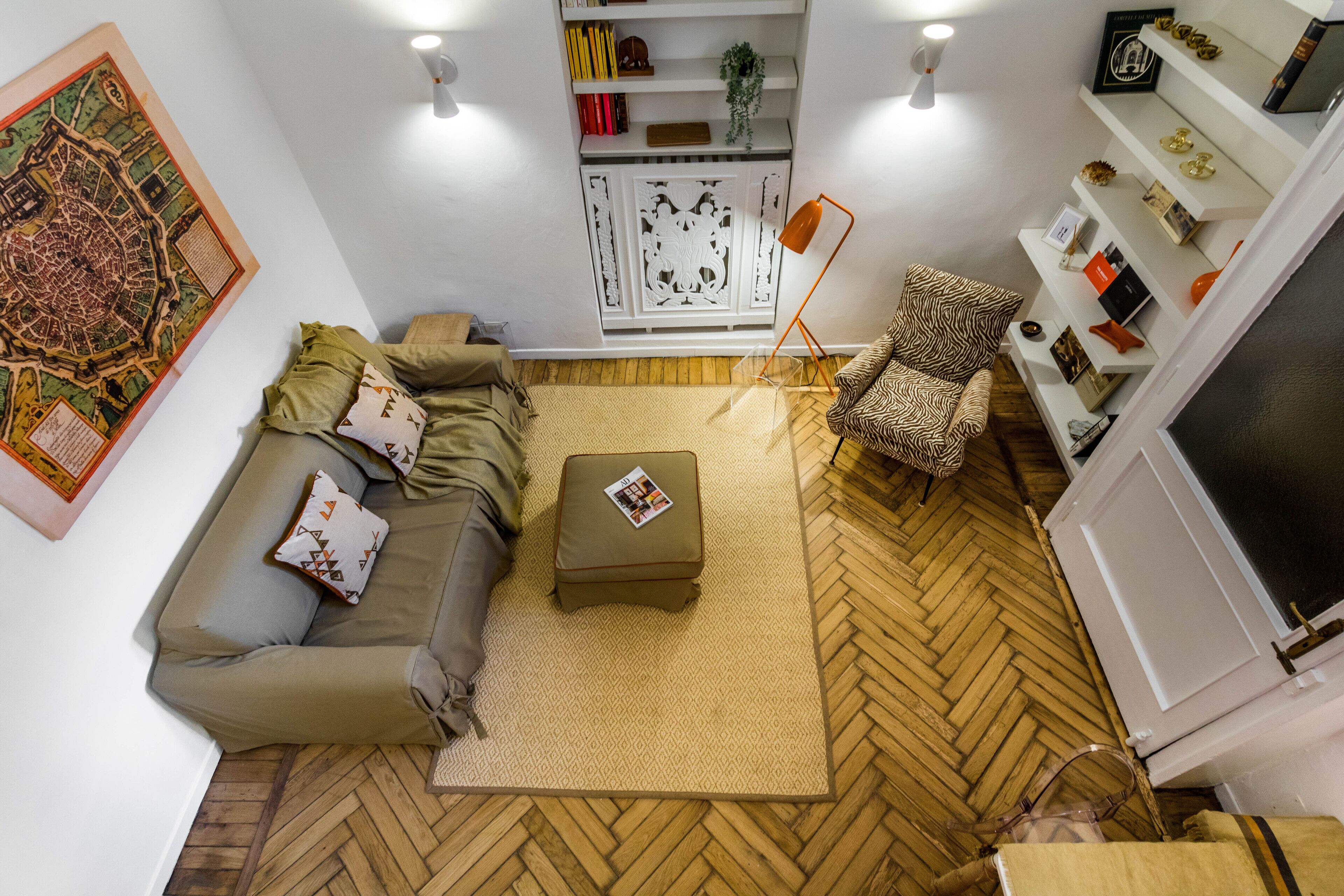 A living room with a herringbone floor, a sofa with patterned cushions, a small ottoman, a patterned armchair, a floor lamp, and a bookshelf with various items.