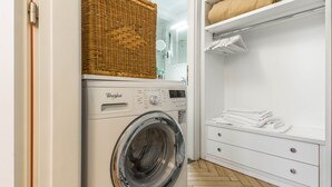 Et lille vaskerum med en frontbetjent Whirlpool vaskemaskine, en fletkurv ovenpå og et hvidt opbevaringsskab med foldede håndklæder.