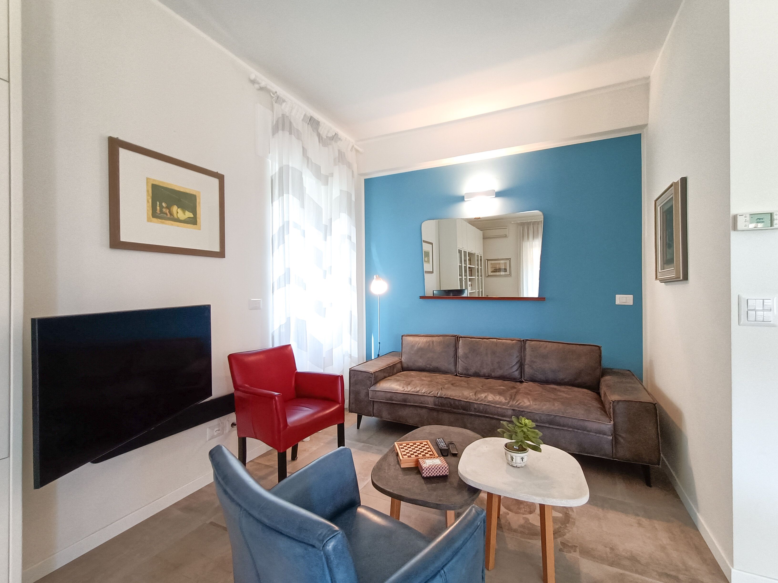 A living room with a flat-screen TV, a red chair, a brown sofa, a blue chair, a small round table with a plant, and a framed picture on the wall.