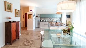 A modern kitchen with teal cabinets, a dining area with a glass table, and a living space with a large window. - JOIVY Bright Flat W/2 Balconies & Sea View (Alassio)