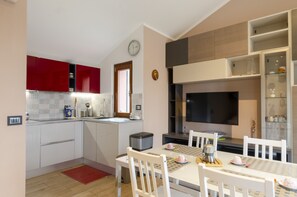 Uma cozinha compacta com armários brancos, uma parede de destaque vermelha, um espaço de refeições com mesa e cadeiras, e uma TV integrada.