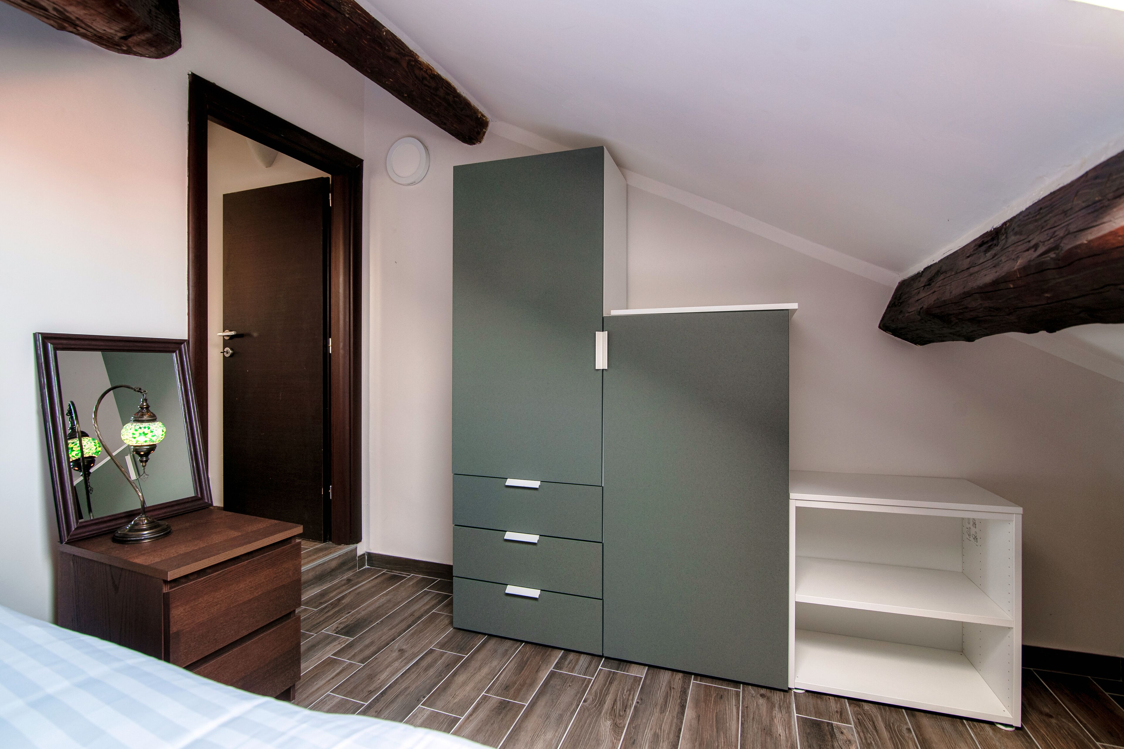 A bedroom with a wooden dresser, a green wardrobe with drawers, and a white shelf unit.