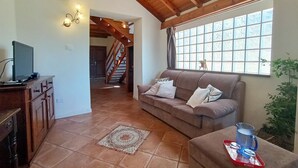 Ein Wohnzimmer mit Holzbalkendecke, einer Couch mit Kissen, einem Fernseher auf einem Holzschrank, einem gemusterten Teppich und einem Fenster mit Blick ins Freie.