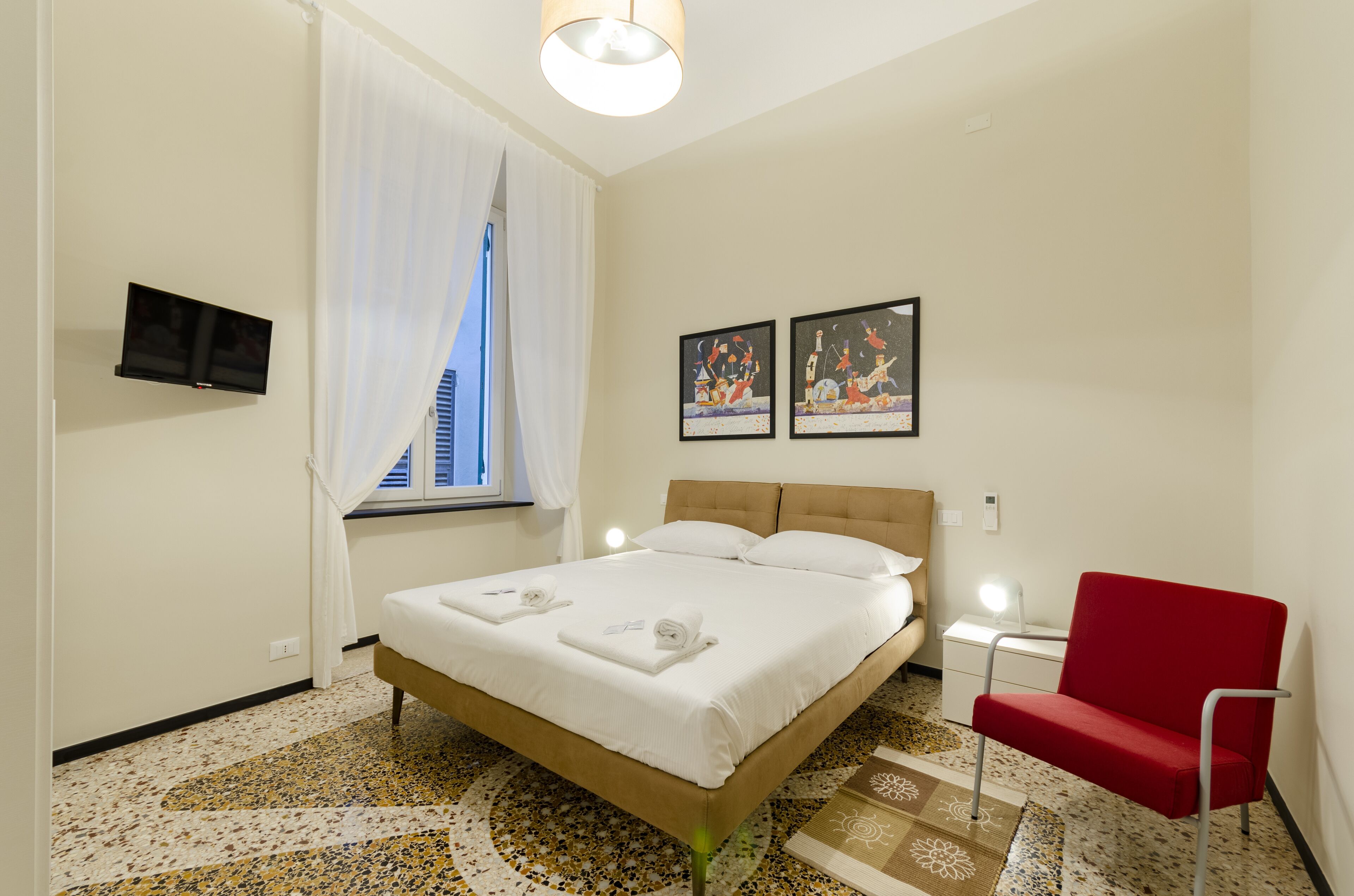 A hotel room with a bed, a red chair, a TV, and artwork on the wall.