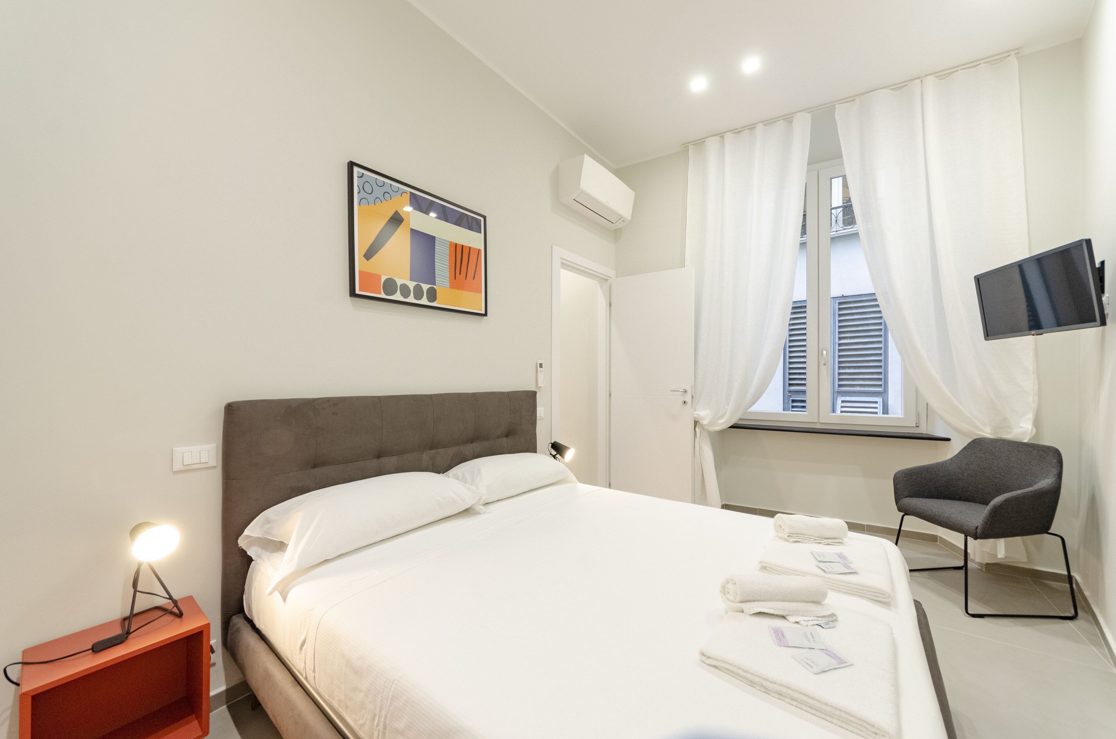 A hotel room with a bed, a chair, a TV, and a painting on the wall.