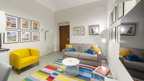 Een moderne woonkamer met een gele fauteuil, een grijze bank met blauwe en gele kussens, een kleurrijk gestreept tapijt en een ronde witte tafel.