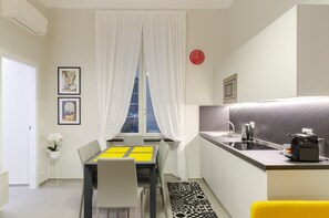 A compact kitchen with a dining area, a window with white curtains, a clock, and a small potted plant. - JOIVY Bright Flat In The Perfect Centre Of Genoa (Genoa)